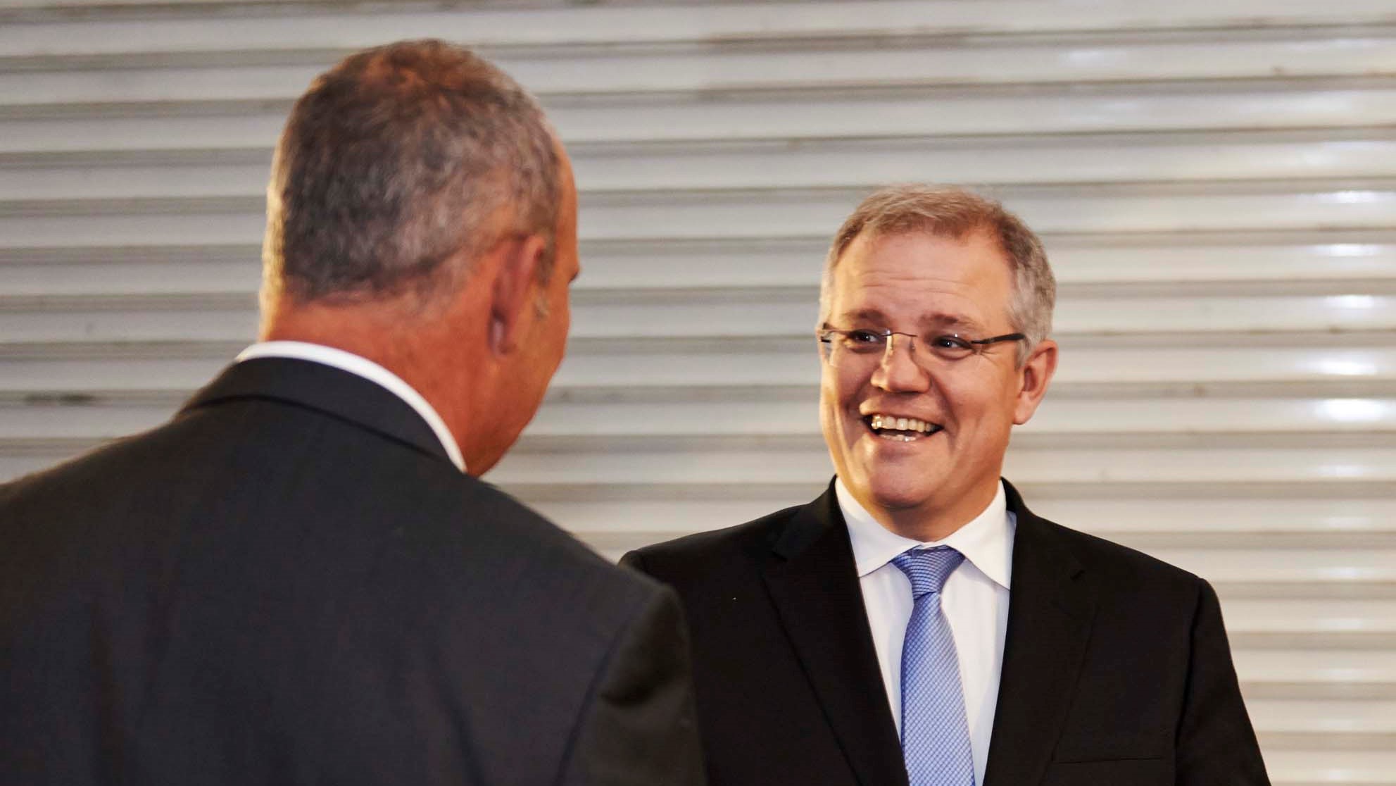 A man in suit and blue tie with glasses smiling and speaking to another man in suit with back facing camera