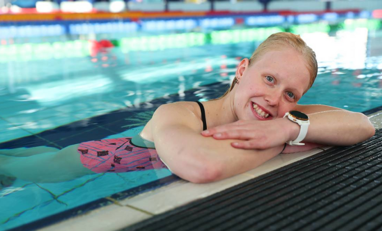 Ash. A young blonde woman swimming in a pool. She is leaning on the edge of the pool and smiling. 