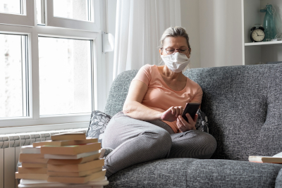 Image of lady wearing a medical mask using her mobile device on a couch.