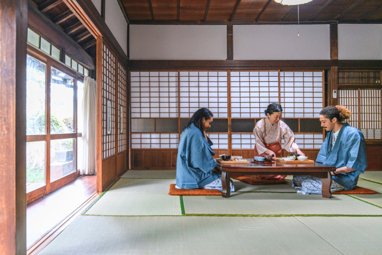Image of three people kneeling at a traditional Japanese table. The room has tatami floor and rice paper walls.