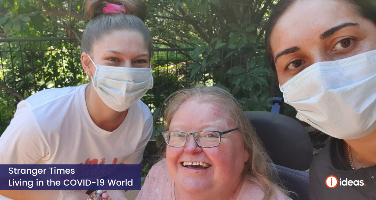 3 women together, in the middle is a woman smiling in a pink top, shes seated in a wheelchair, on either side is another woman, wearing a surgical mask