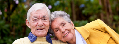 One elderly female is resting their head on the shoulder of another elderly man.