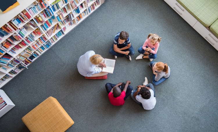 children sitting in a circle listening a story at the library being read by an adult.