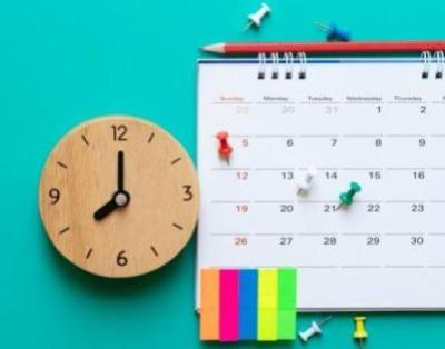 A close up a wooden clock and a calendar with pins and post-it notes.