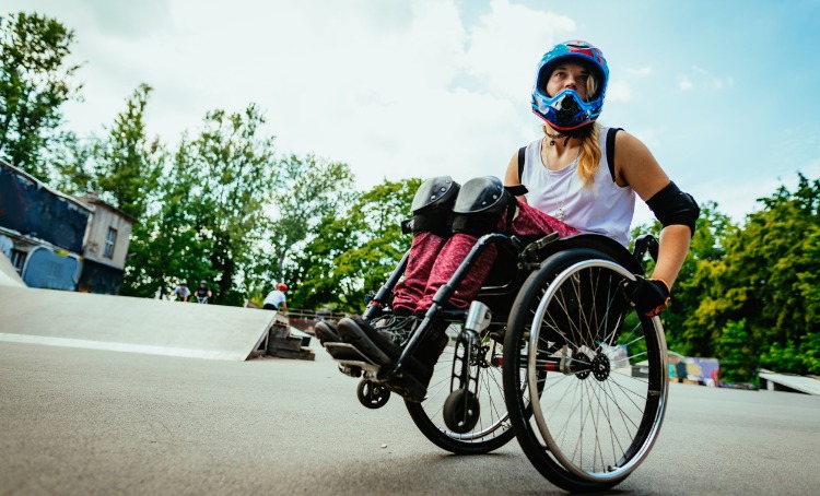 A female in a wheelchairs wears a helmet, knee and elbow guards and gloves and is in a skate park.
