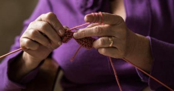 detail of older woman's hands knitting. she is wearing a purple jacket.