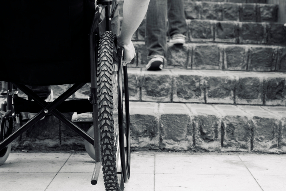 Black and white image of wheelchair in front of a flight of stairs, being unable to go up them.