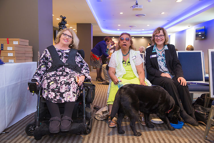 Three women sitting together and smiling. One woman is sitting in a powered wheelchair. A black assistance dog is drinking from a bowl of water at its handler's feet. 