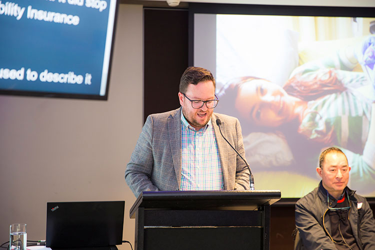 Mr Rick Morton, a man in suit and glasses speaking at a lectern with his head down.