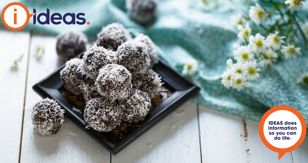 An image of bliss balls stacked on a black plate. They are coated in coconut. A blue floral tablecloth and some flowers are on the right of the photo
