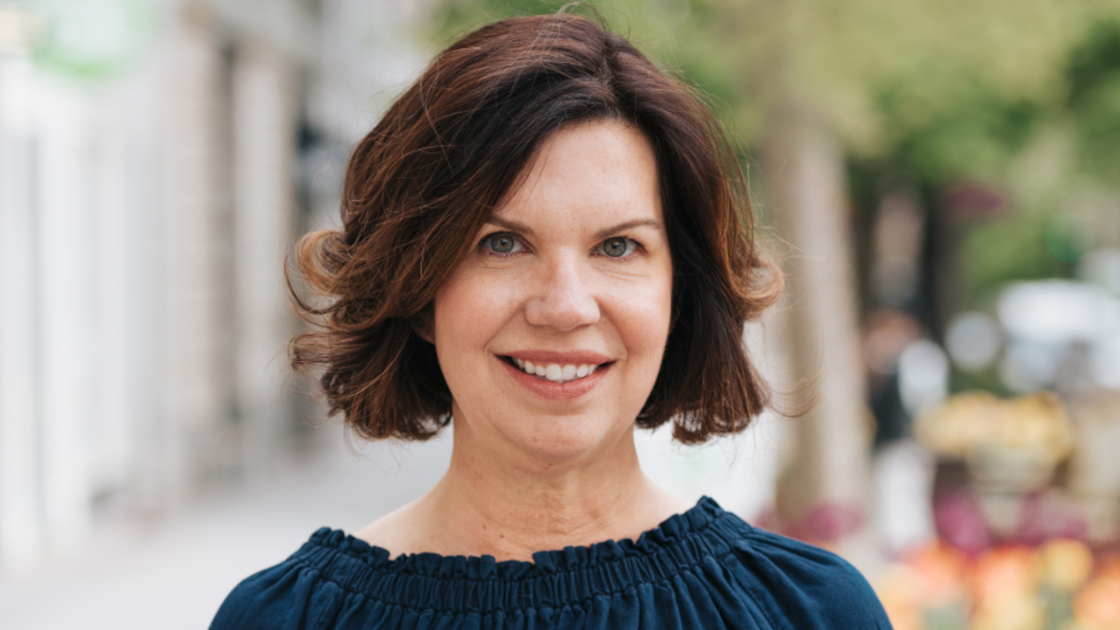 A portrait image of a woman with brown hair and a blue top, she is smiling and looking a the camera. She has an invisible disability.