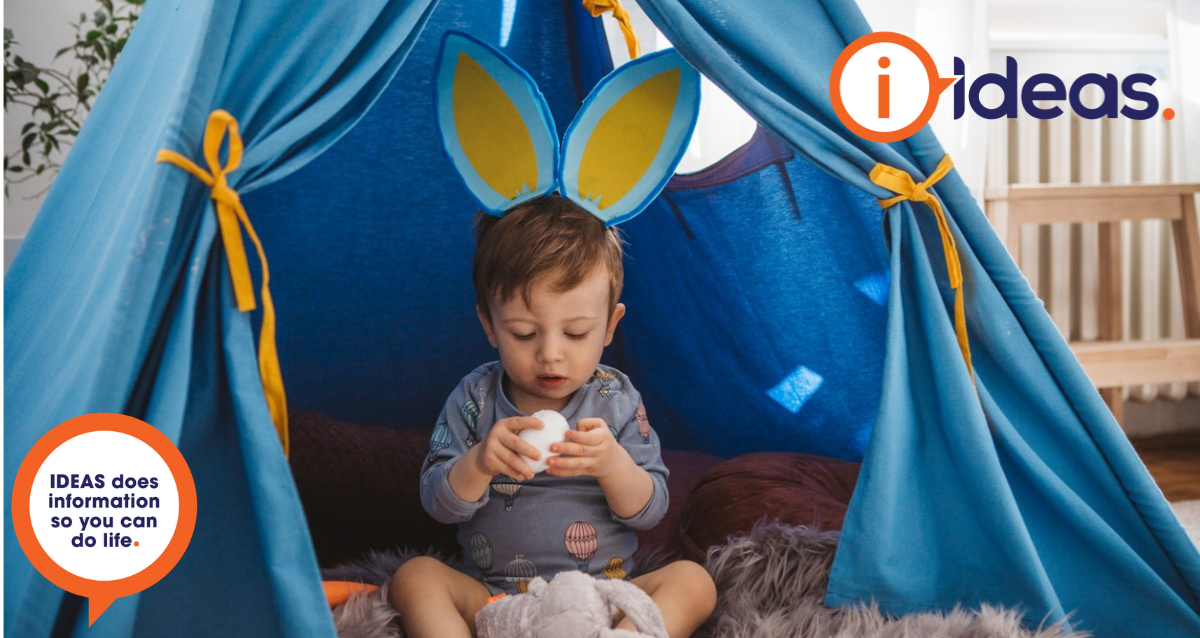 A young boy sits on a fluffy grey lambswool. He wears a pair of Easter Ears in blue and yellow, and has a grey top with hot air balloons on. He is inside a blue indoor play tent. He holds a white Easter Egg which he is looking at and in his lap is a soft toy bunny.