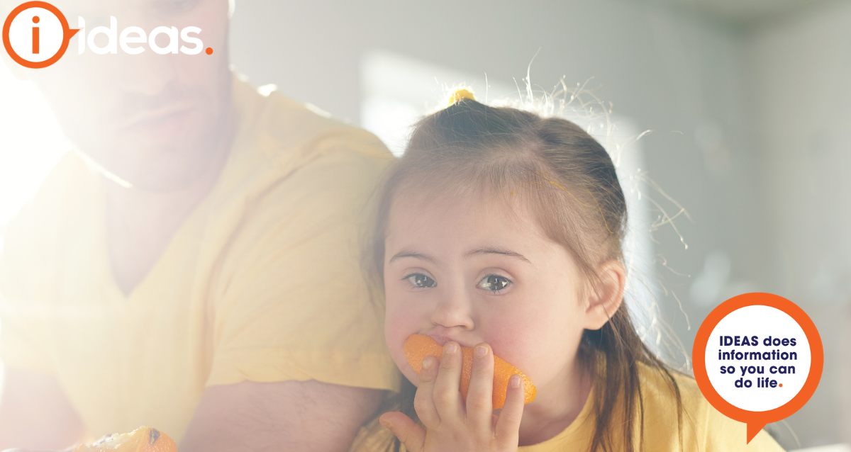 A young girl with down syndrome rests her head against an adult, the girl is eating an orange..