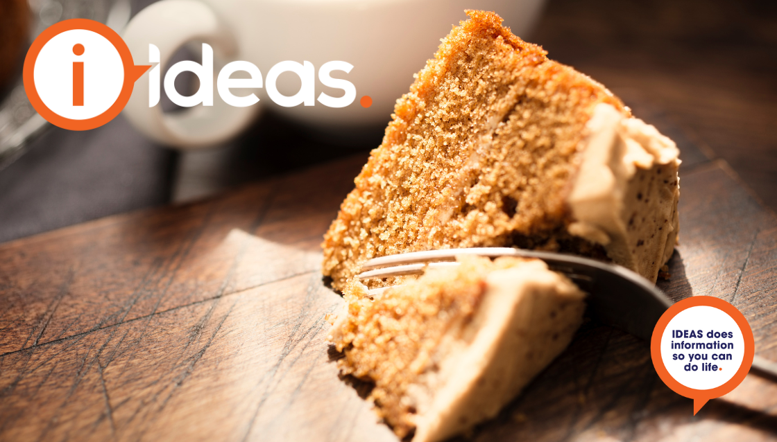 A slice of coffee cake rest on a board, a fork is cutting into the cake. A white cup is in the background.