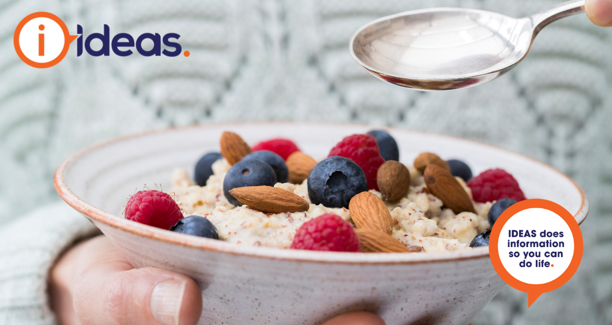 An image of a person in a light grey winter jumper. In one hand they hold a speckled bowl, inside the bowl is porridge, topped with raspberries, blueberries and whole almonds. In the other hand they hold a silver spoon over the bowl.