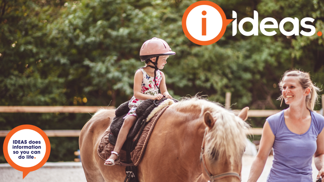 A young girl rides a horse, she wears a pink riding helmet. She smiles at her trainer next to her.