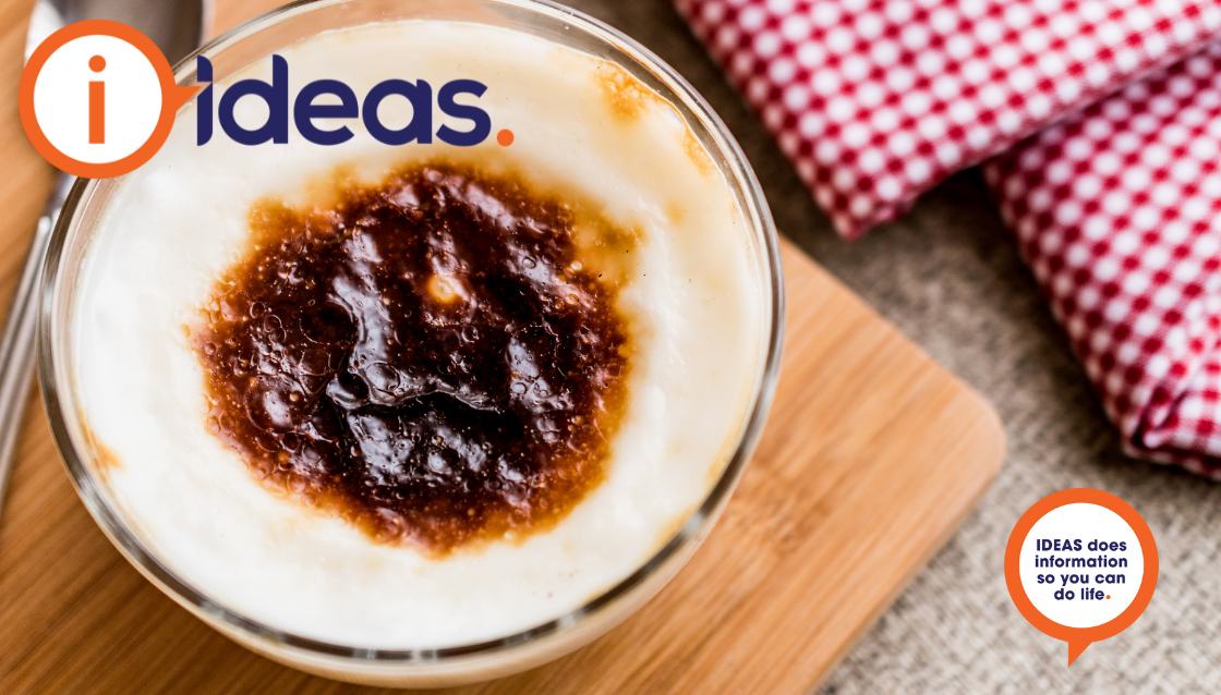 A bowl of rice pudding, with a dark patch on the skin top. ON a bamboo board with a spoon and checked serviette.