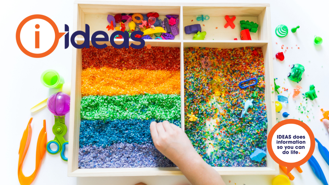 A wooden tray with multicoloured rice within. A childs hand is playing in the tray. There are toys and tools around the tray.