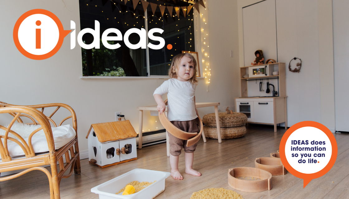 A child plays at home, In the backgroundare fairy lights, bunting, a chair, childs kitchen and toy house. On the floor is a bucket with dried pasta and a cup.