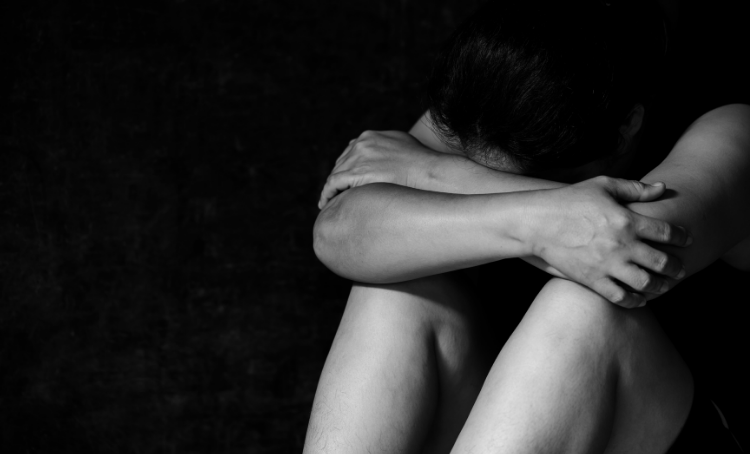 A person with their head bent down, folded arms on their knees. A black and white image.