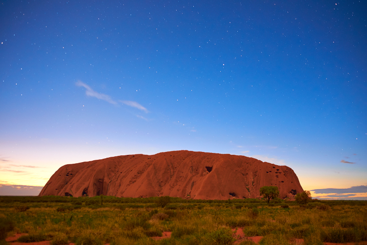A pre-dawn photograph of Uluru with the rock red in the centre and stars in the sky.