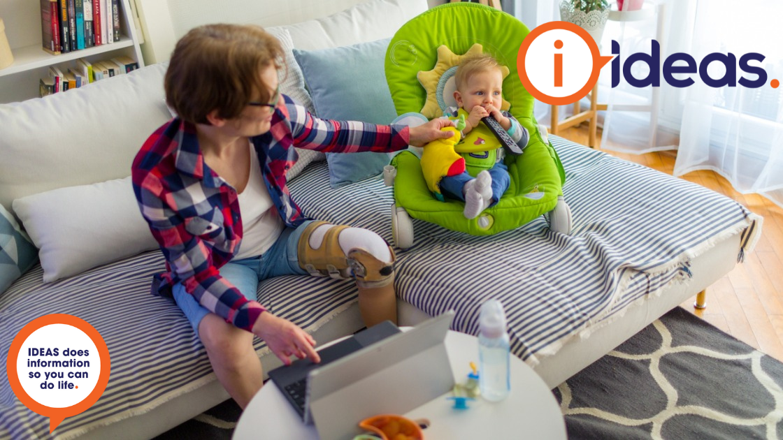 A woman with prosthetic leg works from home on her laptop, her baby is beside her in a baby bouncer