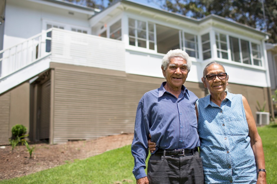 Image of elderly couple smiling with their arms around each other and standing in front of a house.