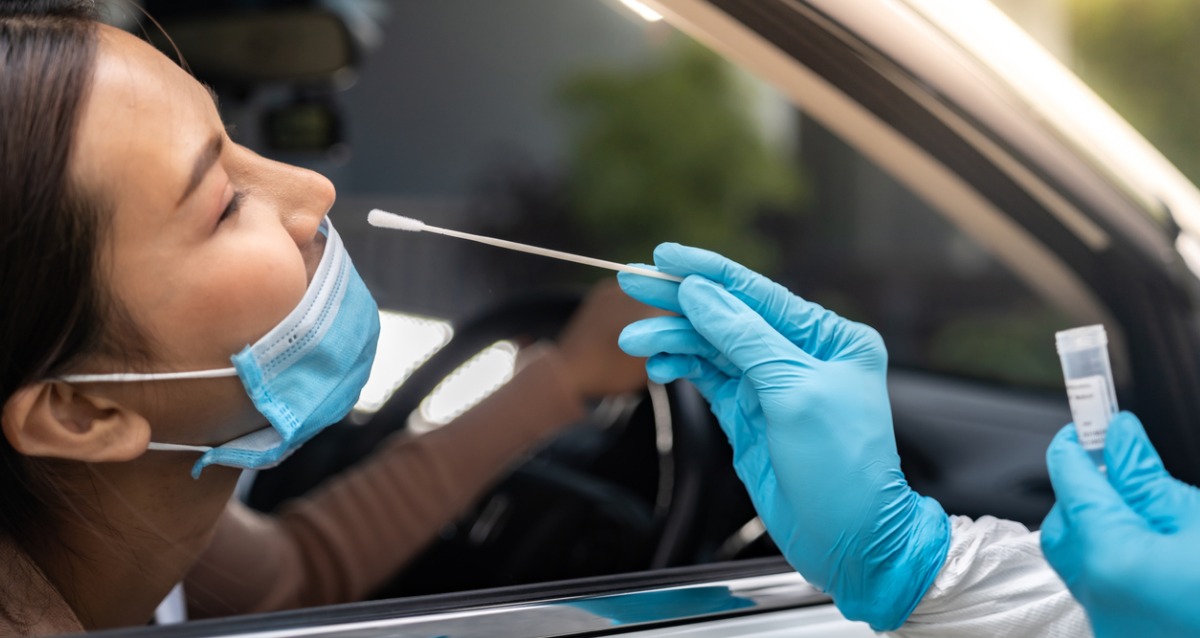 A woman in a car with a medical mask is about to have a covid -19 test. A person off camera is holding a swab and container.