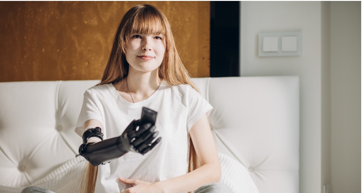 A young woman with long hair, a smile and a prosthetic arm is holding a TV remote and pointing it. 