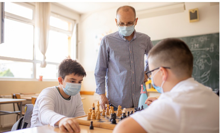 two teenagers playing chess whilst an adult looks on. All are wearing face masks.