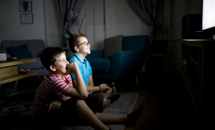 Two boys sit on the floor in a semi-lit room. They face the TV and the light from the TV is illuminating their faces.