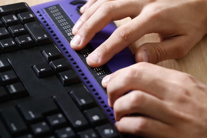 An image of a hands on a braille computer device that is connected to a keyboard. 