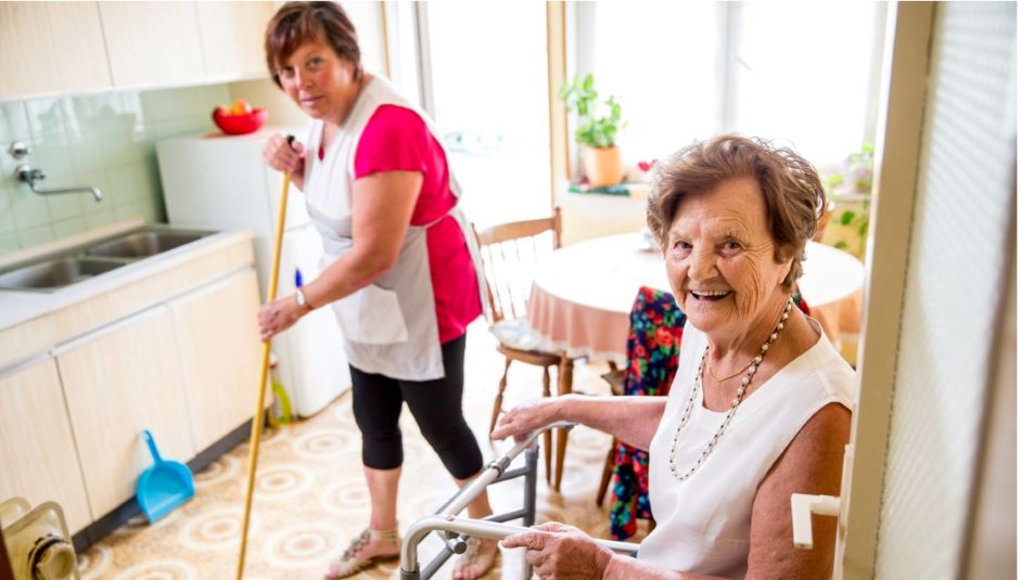 A senior lady sitting and a care giver sweeping the floor.