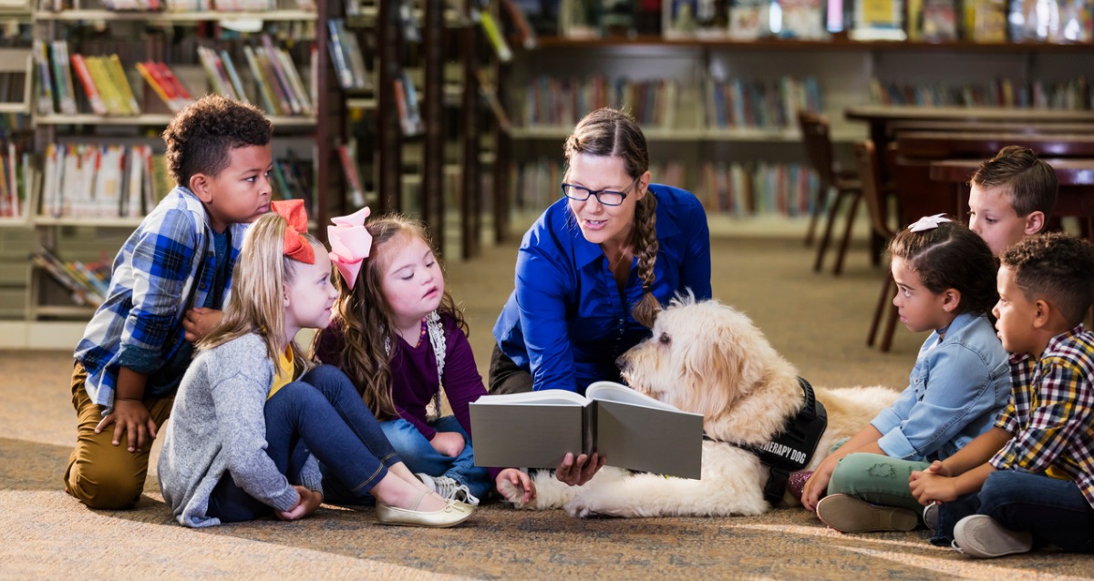children, teacher and assistance dog sitting on the floor in a library reading a book as a group