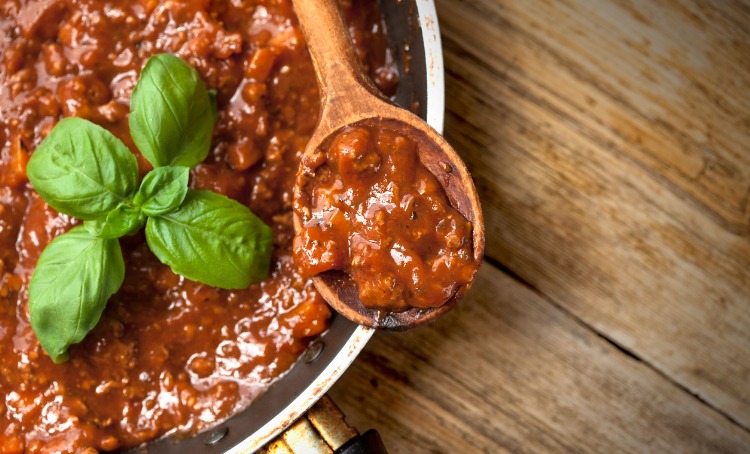 A close up photo of savoury mince in a rich red sauce on a wooden spoon, resting on the side of the cooking pan. A Piece of basil is garnished on top.