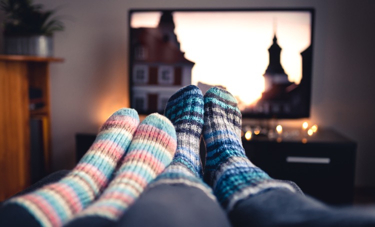 Two pairs of feet wearing warm socks. In the background and out of focus is a TV. Small candles are lit below the TV. Concept photo of watching TV.