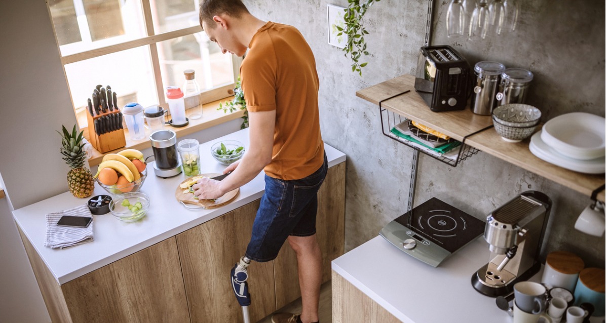 A photograph of a young male with prosthetic leg. He is standing in a kitchen, chopping food. In front of him is a blender to make a smoothie with.