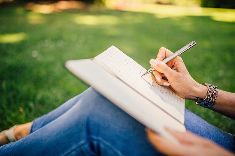A woman sitting outdoors, resting a journal on her legs, writing. Gratitude journal.
