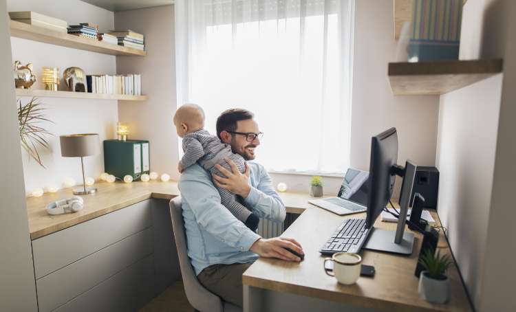 Young father sitting in front of computer trying to work from home while holding his baby boy.