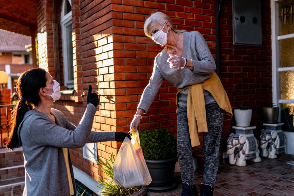 Woman wearing mask and gloves hands a shopping back to an older woman also wearing mask and gloves