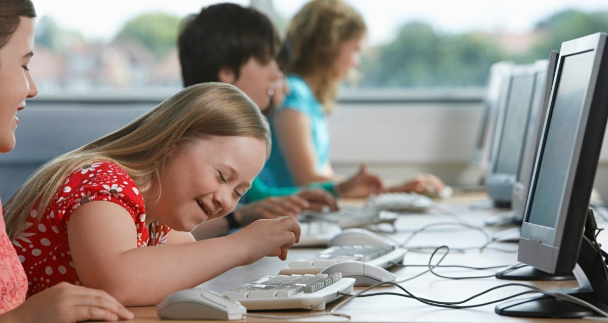 A girl with down Syndrome is smiling and typing on a computer. She is in a room with other children. 