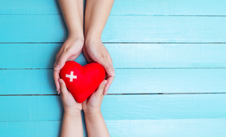 An image with two pairs of hands, holding a red heart between them. The red heart has a white plus on it, first aid symbol.