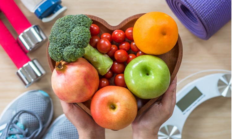 Image of person holding a heart shaped fruit bowl full of colourful fruit and vegetables with fitness equipment in the background.