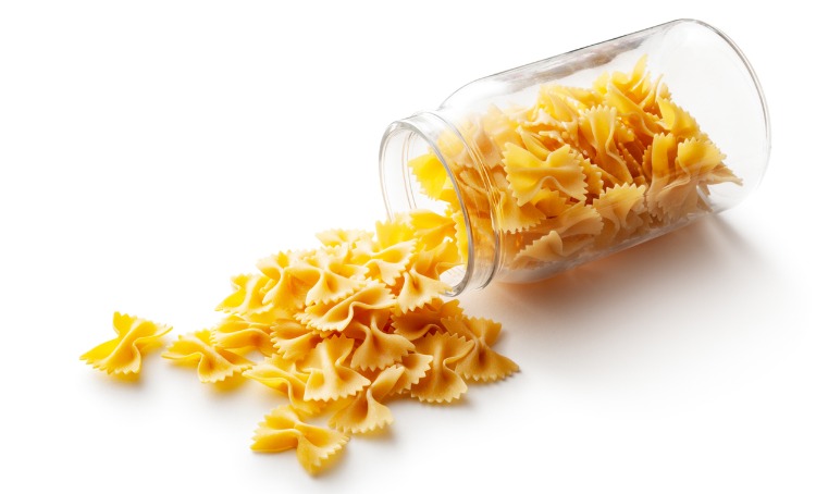 Farfalle fan shaped pasta which is spilling out of a glass jar resting sideways.