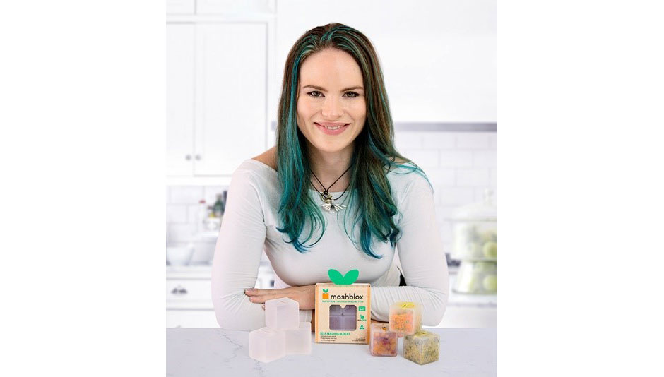 A woman with dark and green hair smiling and sitting behind a Mashblox product display