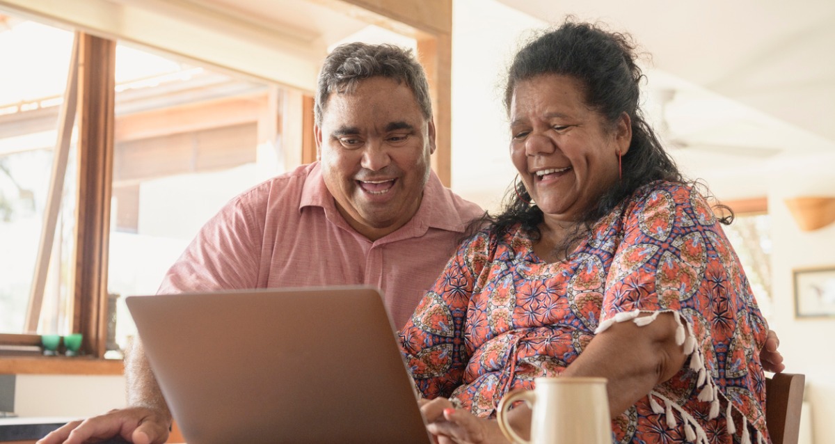 A mature Aboriginal couple sit at a table. In front of them is a laptop, they are both smiling/ laughing at something on the screen. A cup of sits to the left of the woman.