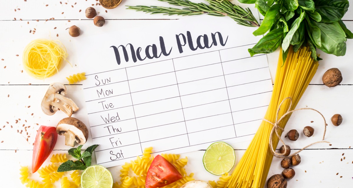 A printed piece of paper with the heading "Meal Plan" has days of the week and columns to place meals. The paper is surrounded by foods like pasta, herbs, vegetables, fruits, nut and spices,
