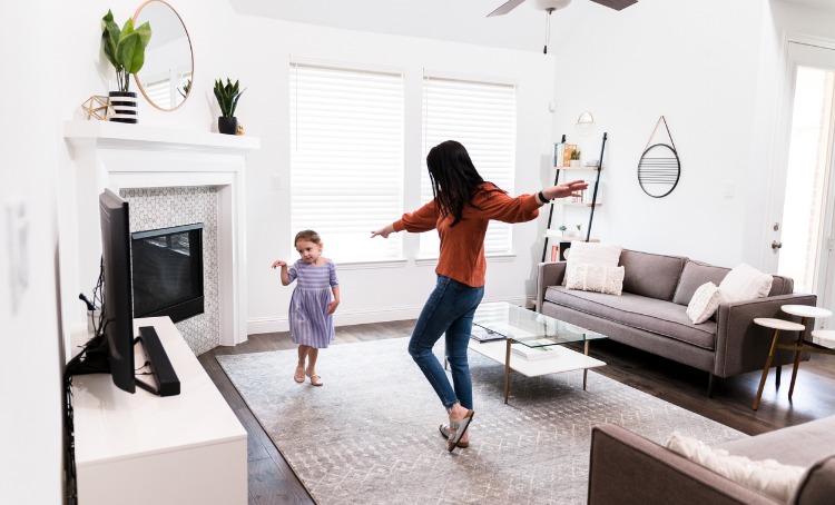 A mother and daughter are dancing to something on the TV. The TV is left of screen. They are in a lounge room with furniture around them in a light and airy space.