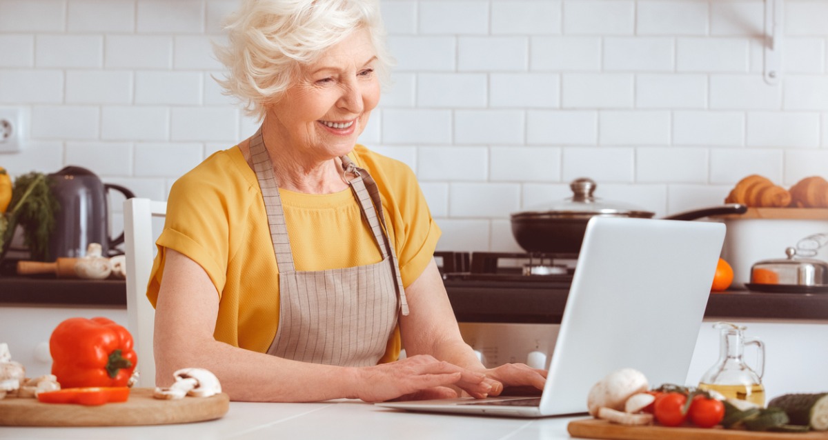 An image of an older woman in a kitchen looking at a laptop. She is wearing an apron and is surrounded by vegetables and cooking utensils.