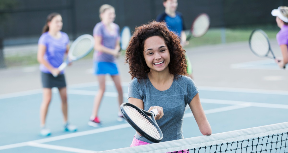 An Image of a young girl with disabilty, standing at a tennis net, holding a tennis racquet.